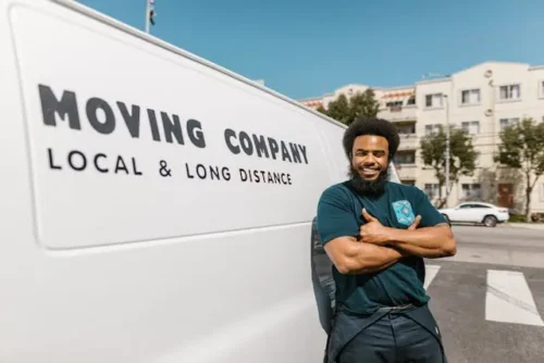 A man in a blue shirt standing proudly next to a moving company van