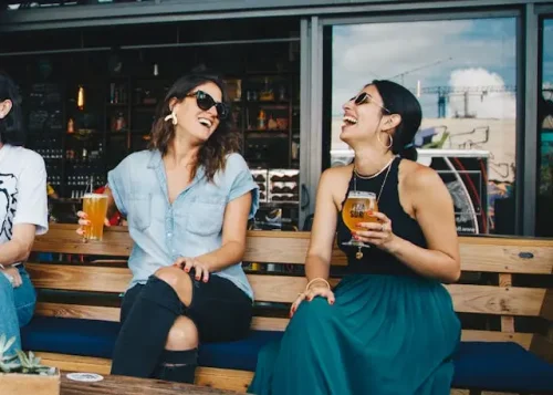 Two women sitting on a wooden bench, laughing and drinking to improve mental health