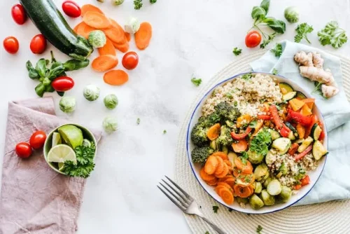 Overhead of a bowl with quinoa and roasted vegetables; sliced carrots, cherry tomatoes, herbs, and limes on a white surface