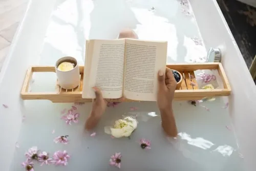 Person soaking in a milky bath, holding an open book on a wooden tray with flowers and a mug