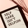 Letter board reading “SELF CARE ISN’T SELFISH” on a pink surface with palm shadow, representing wellness rituals to reset